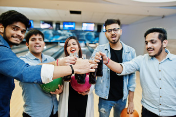 Group of five south asian peoples having rest and fun at bowling club. Clinking cold soda drinks from glass bottles and bowling balls at hands.