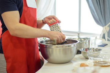 man in a red apron in the kitchen rub potatoes on a grater