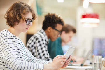 Serious thoughtful student girl in glasses sitting at table in university and using digital tablet while searching for information on internet 