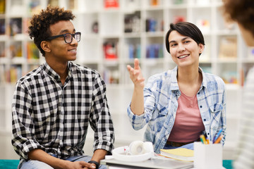 Cheerful creative interracial students in casual clothing sitting in library and discussing ideas while working together on project 
