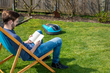 junger Mann im Liegestuhl neben Roboterrasenmäher im Garten