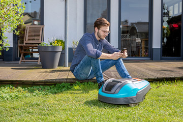 junger Mann sitzt mit Handy hinter Roboterrasenm&auml;her