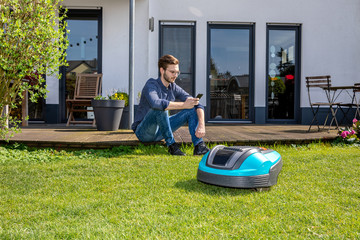 junger Mann sitzt mit Handy hinter Roboterrasenmäher, Garten