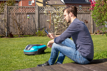 junger Mann mit Handy neben Mähroboter im Garten