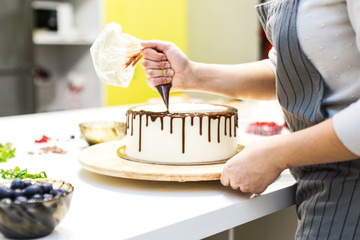 A confectioner squeezes liquid chocolate from a pastry bag onto a white cream biscuit cake on a...