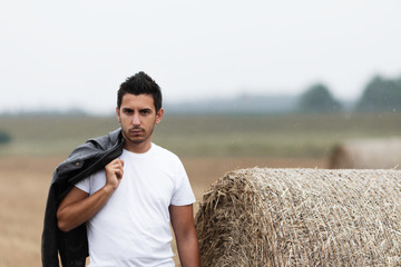 A handsome young brunette man stands in a field near a haystack.
