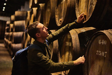Young man tourist  walk in old aged traditional wooden barrels with wine in a vault lined up in...