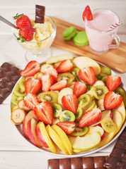 Chocolate bars and fruit sliced ​​from strawberries, apples, bananas and kiwi on a wooden white background.