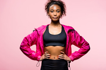 attractive african american sportswoman in windbreaker looking at camera and posing with hands akimbo isolated on pink © LIGHTFIELD STUDIOS