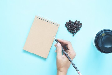 Creative flat lay of heart shape coffee beans and woman hand holding pencil with notebook on blue colour background