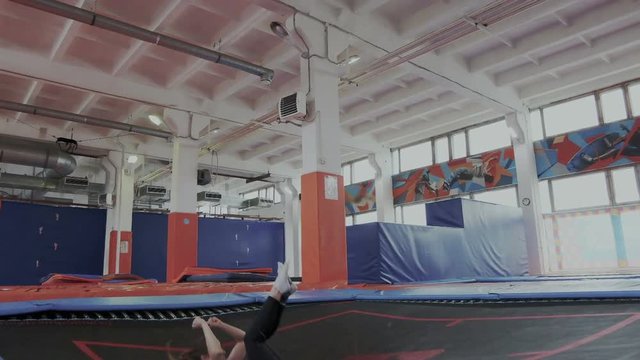 Young 20s athlete girl shows acrobatic stunts jumping on a trampoline. She slowly levitates in the air and groups her body and falls to the surface of the trampoline