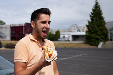 Handsome young brunette man eating hot dog in the parking lot near the gas station.