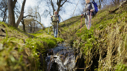 Fototapeta premium Fresh breathe. Aged family couple of man and woman in tourist outfit walking at green lawn near by trees and creek in sunny day. Concept of tourism, healthy lifestyle, relaxation and togetherness.