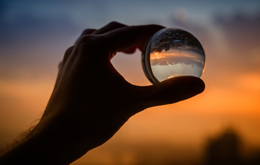 Hand holds glass ball which reflects sunset sky over city.
