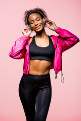 Obraz premium beautiful smiling african american sportswoman in headphones listening music isolated on pink