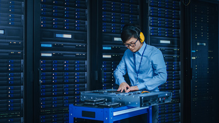 In the Modern Data Center: IT Technician Wearing Protective Headphones Working with Server Racks,...