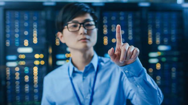 In the Modern Data Center: Portrait of Asian IT Engineer Working With Touch Screen Device, Doing Touching Gesture. In the Background Working Server Racks with Blinking LED Lights. - Powered by Adobe