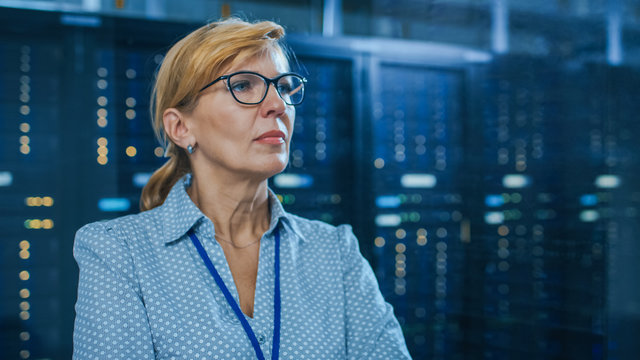 Portrait of a Beautiful Senior Female IT Technician Standing In Modern Data Center. In the Background Working Server Racks with Blinking LED Lights.
