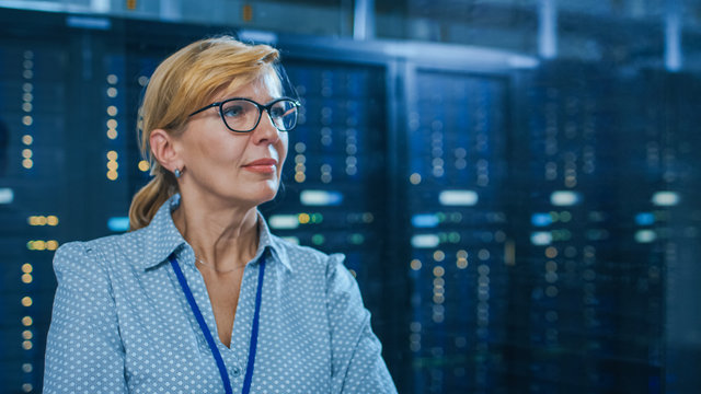 Portrait of a Beautiful Senior Female IT Technician Standing In Modern Data Center. In the Background Working Server Racks with Blinking LED Lights. - Powered by Adobe