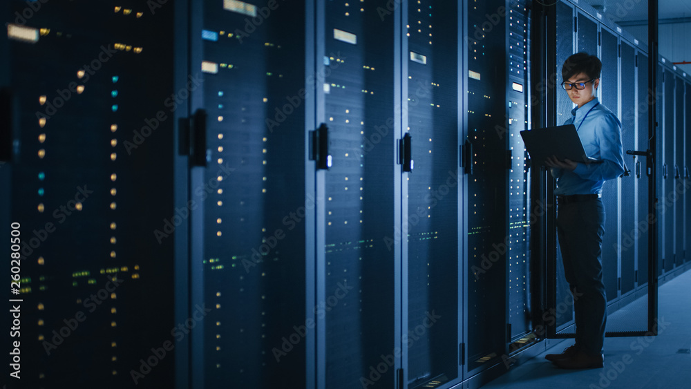 Poster in dark data center: male it specialist stands beside the row of operational server racks, uses lapt