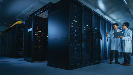 In the Data Center: Two IT Specialists Wearing White Lab Coats, Have Discussion about Working of...