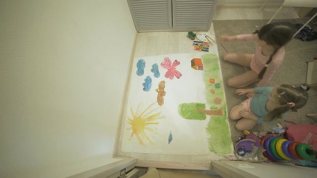 two girls paint with watercolor paints and brushes on a large drawing paper on the floor , children's joint creativity