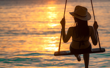 Silhouette woman wear bikini and straw hat swing the swings at the beach on summer vacation at...