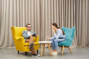 Full length portrait of young woman talking to mature psychologist in therapy session sitting on...