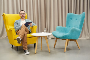 Full length portrait of mature psychologist holding clipboard  posing looking at camera while  sitting on design chair against drapery, copy space
