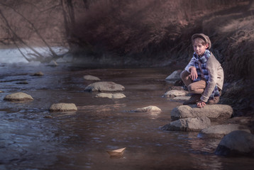 boy sits on a stone in a creek and catches a paper boat, vintage photo, launch a craft craft in the river