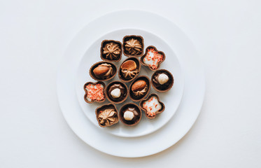 Beautiful chocolates of different shapes and fillings lie in a white plate against the grey background paper. Copy space.