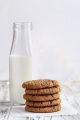 Stack of fresh homemade oatmeal cookies with a bottle of milk on a white table against a white background.