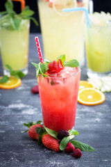 Glass of cold strawberry drink with ice cubes, mint leaves. Berries, orange slices on textured wooden table. Lemonade jar and glasses at the background out of focus.