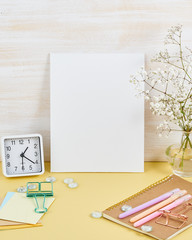 Mockup with blank white frame on yellow table against wooden wall, alarm, flower in vaze, vertical