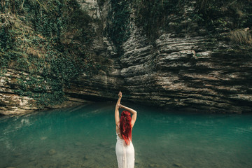 Attractive girl with long red hair is standing with his back in blue water near a rocks.