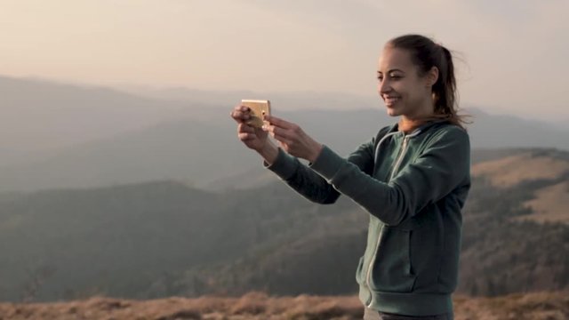 young woman standing on mountain ridge and enjoy the view at sunset and taking a photo using phone
