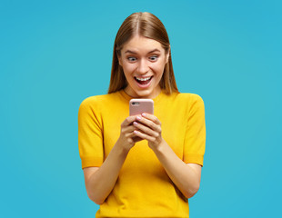 Stunned girl having widely opened eyes and mouth, looks at the phone. Photo of girl in yellow...