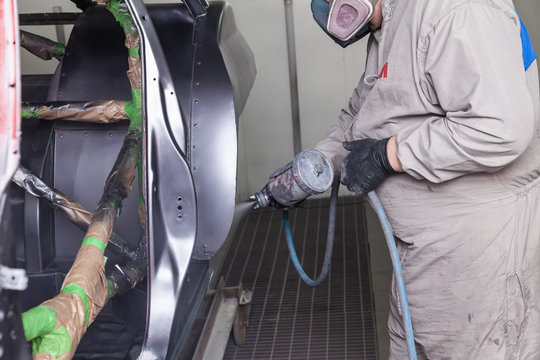 A man in protective overalls and a mask holds a spray bottle in his hand and sprays black paint onto the frame of the car body after an accident during a repair in a vehicle restoration workshop
