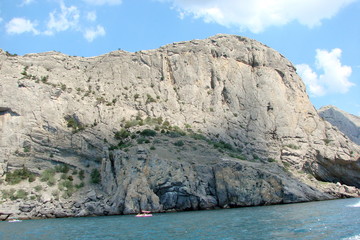 Panorama from the ship's side of the naked coastal rocks under the sun's rays against the background of a cloudy blue summer sky.