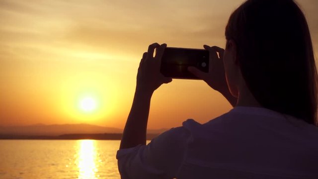 Dark silhouette of young woman taking photos on cellphone at sunset on lake while enjoying quiet moment in nature. Carefree female figure making pictures on mobile phone at golden hour in slow motion