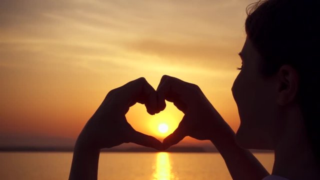 Dark silhouette of young woman at sunset at sea making heart gesture with fingers. View from behind of female figure at golden hour catching sun in hands folded in heart shape