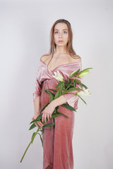 shy caucasian girl with big eyes stands in a velvet pink dress and holds a white Lily in her hand as a symbol of innocence and purity. young woman posing on white studio background.