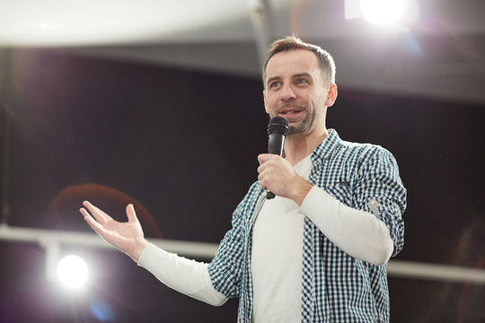 Waist up portrait of mature man giving speech standing on stage in spotlight and speaking to microphone, copy space