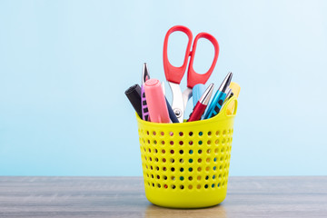 Colorful pens and orange scissors are in the green plastic basket and put on wooden table isolated on blue pastel background.