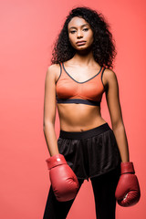 beautiful african american sportswoman in boxing gloves looking at camera and posing isolated on coral