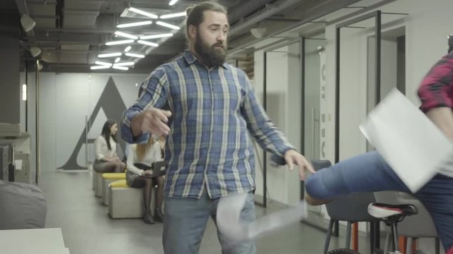 Bearded man studying papers in the office. Young colleague riding the bike in the office and pushes bearded guy in the shoulder, papers fall down. Bike rider apologizing and collecting documents