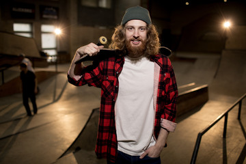 Waist up portrait of bearded man holding skateboard smiling at camera standing in extreme park, shot with flash © Seventyfour