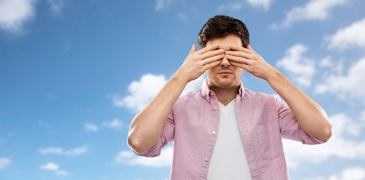 vision and people concept - man closing his eyes by hands over blue sky and clouds background