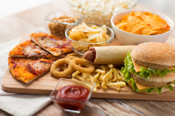 fast food and unhealthy eating concept - close up of double hamburger or cheeseburger, deep-fried squid rings, french fries, pizza and chrisps on wooden board