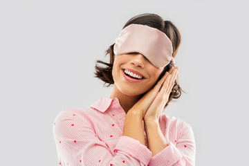 people and bedtime concept - happy young woman in pajama and eye sleeping mask over grey background